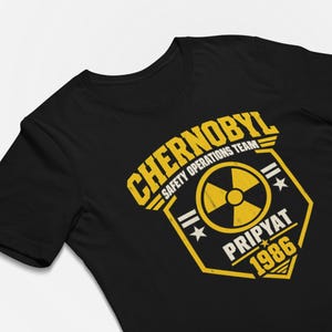 Könnte beinhalten: Ein schwarzes Kurzarm-T-Shirt mit gelbem Grafikdruck. Das Design zeigt ein Strahlensymbol in einer Schildform mit dem Text CHERNOBYL SAFETY OPERATIONS TEAM, PRIPYAT und 1986 in einer fetten, abgenutzten serifenlosen Schriftart.