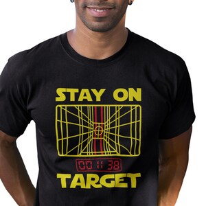 May include: A black short-sleeved t-shirt featuring a yellow graphic of a targeting computer display with the text STAY ON TARGET in yellow above and below the graphic, and a red digital timer reading 00 11 38 centered below the display.
