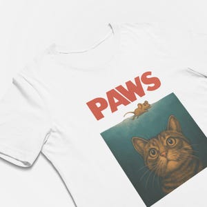 May include: White t-shirt with the word "PAWS" in red block letters. Below the text is a graphic of a cat looking up at a mouse. The cat is brown and tan with large eyes, and the mouse is brown.