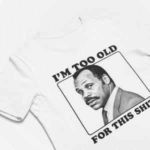 May include: A white short-sleeved t-shirt featuring a black and white graphic print of a man in a suit within a rectangular frame, accompanied by the bold black text "I'M TOO OLD FOR THIS SHIT." printed above and below the image.