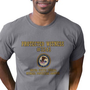 May include: A gray t-shirt with the text "PROTECTED WITNESS #3243" and a seal of the United States Department of Justice with the text "UNITED STATES FEDERAL WITNESS PROTECTION PROGRAM".