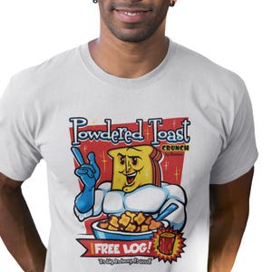May include: A white t-shirt featuring a cartoon illustration of a superhero made of powdered toast. The illustration is in the style of vintage cereal box art. The text on the shirt reads "Powdered Toast Crunch" and "Free Log! It's big, it's heavy, it's wood!"