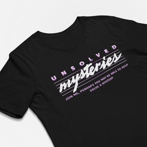 May include: A black short-sleeved t-shirt featuring the text "UNSOLVED mysteries" in a white and light purple gradient font, accompanied by the text "JOIN ME, PERHAPS YOU MAY BE ABLE TO HELP SOLVE A MYSTERY" printed in white below the main title.