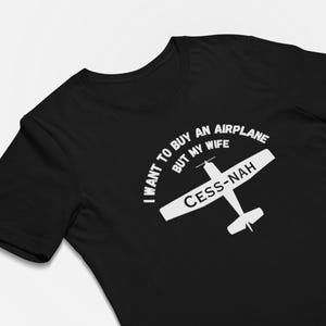 May include: A black short-sleeved t-shirt featuring a white graphic of a small airplane. The text printed in white reads: I WANT TO BUY AN AIRPLANE BUT MY WIFE CESS-NAH. The design is centered on the chest of the garment, which has a crew neckline.
