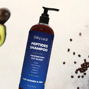 May include: A brown bottle of Shiny Leaf Peptides Shampoo with a black pump, featuring a blue label with white text. Ingredients include Biotin and Caffeine. The image also shows avocados, coffee beans, and a bowl of powder.