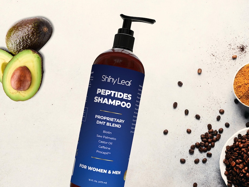 May include: A brown bottle of Shiny Leaf Peptides Shampoo with a black pump, featuring a blue label with white text. Ingredients include Biotin and Caffeine. The image also shows avocados, coffee beans, and a bowl of powder.