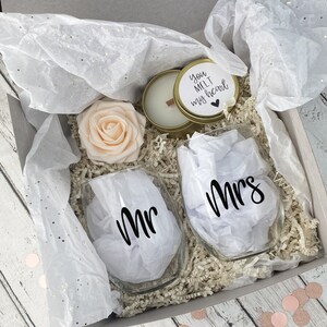 Puede incluir: Una caja de regalo llena de una vela blanca con tapa dorada, dos copas de vino transparentes con las palabras "Mr." y "Mrs." en ellas, una rosa melocot&oacute;n y papel de seda blanco. La tapa de la vela tiene una pegatina dorada con el texto "You melt my heart".