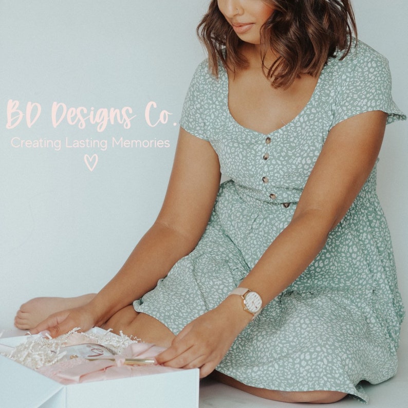 Puede incluir: Una mujer con un vestido verde floral con botones en la parte delantera, se sienta en el suelo y abre una caja de regalo blanca. La caja est&aacute; llena de papel de seda rosa y un bol&iacute;grafo dorado. El texto "BDD Designs Co. Creating Lasting Memories" es visible en la esquina superior izquierda de la imagen.