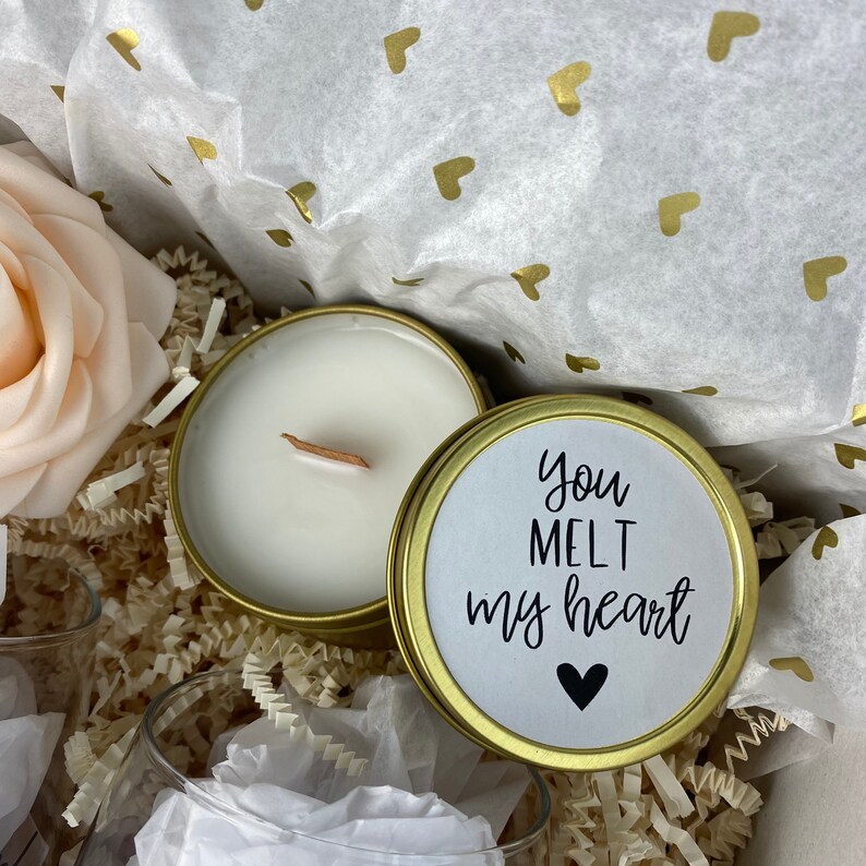 Puede incluir: Una lata de vela dorada con una vela blanca en su interior y una tapa dorada con una etiqueta blanca que dice "you melt my heart" con un s&iacute;mbolo de coraz&oacute;n negro.