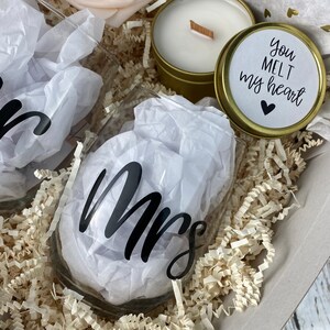 Puede incluir: Una caja de regalo llena de papel triturado, dos copas de vino con la palabra "Mrs." en ellas, una vela en una lata dorada y una peque&ntilde;a lata dorada con las palabras "Me derrites el coraz&oacute;n" en ella.