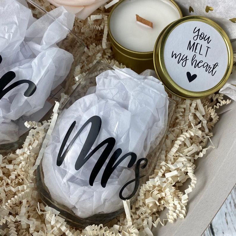 Puede incluir: Una caja de regalo llena de papel triturado, dos copas de vino con la palabra "Mrs." en ellas, una vela en una lata dorada y una peque&ntilde;a lata dorada con las palabras "Me derrites el coraz&oacute;n" en ella.