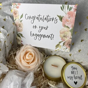 Puede incluir: Una caja de regalo llena de una vela blanca en una lata dorada, una rosa artificial de color melocot&oacute;n y una tarjeta que dice "&iexcl;Felicidades por su compromiso!" La lata de la vela tiene una etiqueta que dice "Me derrites el coraz&oacute;n" con un coraz&oacute;n negro.