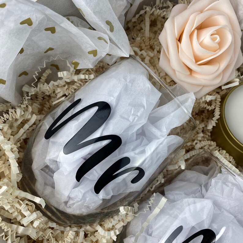 Puede incluir: Dos copas de vino de vidrio transparente con letras de vinilo negro que dicen "Mr" y "Mrs". Las copas est&aacute;n en una caja de regalo con papel de seda blanco y una rosa blanca.