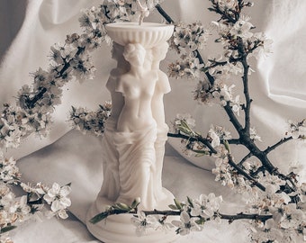 May include: White candle in the shape of a classical sculpture of three women standing together. The candle is surrounded by white blossoms.