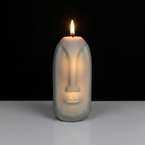 May include: A gray candle shaped like a face with a lit wick. The candle has a nose, lips, and eye indentations. The background is black, and the candle sits on a white surface. The flame is yellow and orange.