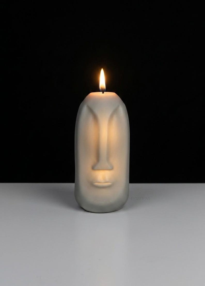 May include: A gray candle shaped like a face with a lit wick. The candle has a nose, lips, and eye indentations. The background is black, and the candle sits on a white surface. The flame is yellow and orange.