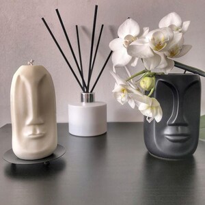 May include: A cream-colored candle and a black vase, both shaped like faces, sit on a dark surface. A white diffuser and a sprig of white orchids complete the arrangement. The candle is on a small, round, black stand.
