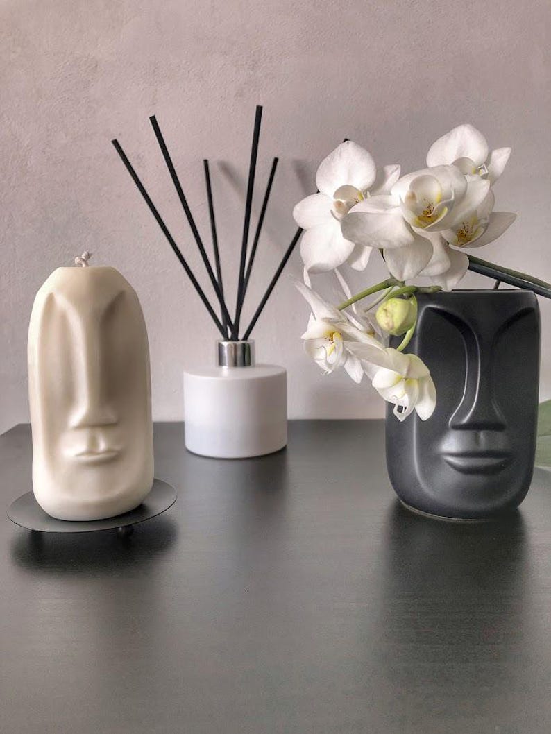 May include: A cream-colored candle and a black vase, both shaped like faces, sit on a dark surface. A white diffuser and a sprig of white orchids complete the arrangement. The candle is on a small, round, black stand.