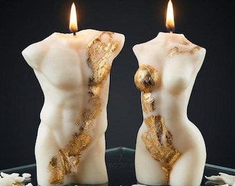 May include: Two off-white torso-shaped candles, one masculine and one feminine, both lit with flickering flames. The wax figures are decorated with irregular patches of metallic gold leaf applied to the surface, creating a textured appearance.