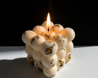 May include: A lit, ivory-colored bubble candle with gold leaf accents. The candle is cube-shaped, with each bubble-like sphere adorned with gold flecks. A single flame burns atop the candle, with a wisp of smoke rising. The background is black and white.