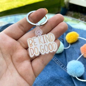 May include: A white acrylic keychain with the text "Be Kind Do Good" in gold lettering. The keychain is attached to a silver metal ring.