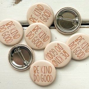 Puede incluir: Seis botones redondos de color beige con el texto "Be Kind Do Good" en un degradado de naranja, rosa y marrón. Los botones tienen una parte posterior plateada con un pasador de seguridad.