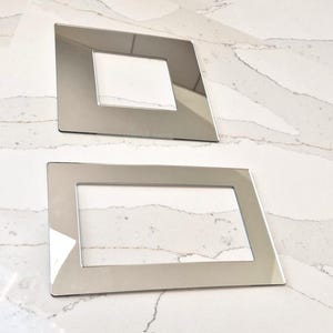May include: Two silver-coloured wall plate covers with a rectangular opening in the centre. The covers are made of a reflective material and have a smooth, polished finish.