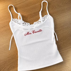 May include: White ribbed vest top with lace trim and spaghetti straps. The text "Mrs. Cavillo" is embroidered in red script across the front. The top has a tie detail at the neckline.