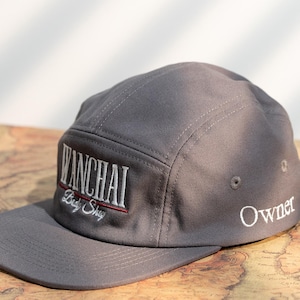 Puede incluir: Gorra gris de cinco paneles con visera plana. El panel frontal presenta el texto bordado "WANCHAI" en blanco, con "Lady Shop" debajo en letras más pequeñas. Una línea roja acentúa el texto. La palabra "Owner" está bordada en el lateral.