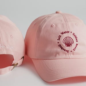 May include: Pink baseball cap with a white embroidered design on the back and a red embroidered design of a seashell with the text "Salt Water Golden Beach Seagull" on the front.