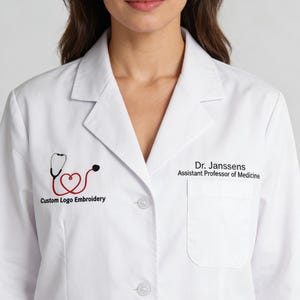 Op de afbeelding: Witte laboratoriumjas met een rood-zwart stethoscooplogo geborduurd op de linkerborst. De tekst "Custom Logo Embroidery" staat onder het logo. De rechterborst heeft een zak en de tekst "Dr. Janssens, Assistant Professor of Medicine".