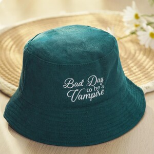 May include: A teal corduroy bucket hat with the text "Bad Day to be a Vampire" embroidered in white script. The hat is sitting on a woven straw surface.