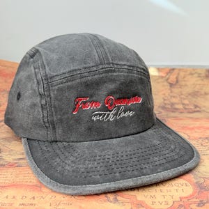 May include: A gray, five-panel cap with a flat brim. The cap features red embroidered text that reads "From Oceanside with love." The hat is made of a textured fabric and has a classic, casual style.