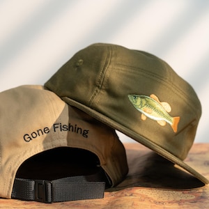 May include: Two canvas caps, one khaki and one olive green, are displayed. The khaki cap has "Gone Fishing" embroidered on the back. The olive green cap features a fish graphic. Both have a black adjustable strap.