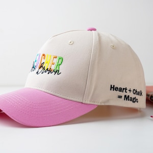 Op de afbeelding: Beige baseballpet met een roze klep. De pet heeft de tekst "Teacher" in kleurrijk borduursel en "Heart + Chalk = Magic" aan de zijkant. Een modieus accessoire.