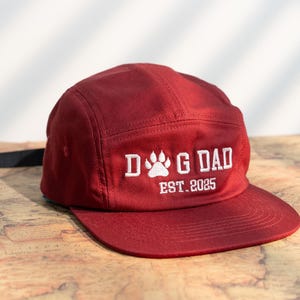 Może przedstawiać: Bordowa czapka typu five-panel z płaskim daszkiem. Na czapce znajduje się biały haft z napisem "DOG DAD" z odciskiem łapy i "EST. 2025". Czapka wykonana jest z wytrzymałego materiału i posiada czarny, regulowany pasek.