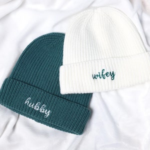 Op de afbeelding: Twee gebreide mutsen, één teal en één witte, liggen op witte stof. De teal muts heeft het woord "hubby" in wit geborduurd, terwijl de witte muts "wifey" in teal heeft geborduurd. De mutsen zijn een bijpassende set.