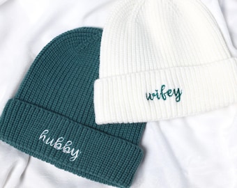 May include: Two knitted beanies, one teal and one white, rest on a white fabric. The teal beanie has the word "hubby" embroidered in white, while the white beanie has "wifey" embroidered in teal. The beanies are a matching set.