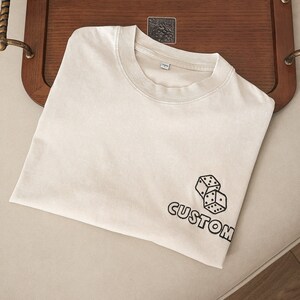 Peut inclure: Un t-shirt beige avec un motif noir de deux dés et le mot "CUSTOM" sur la manche.