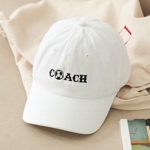 Puede incluir: Gorra de béisbol blanca con un balón de fútbol bordado en negro y la palabra "COACH" en la parte delantera.