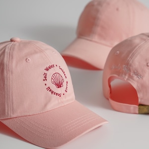 Op de afbeelding: Drie lichtroze baseballpetten. De voorste pet heeft een geborduurd schelpmotief met de woorden "Salt Water, Golden Beach, Seashell". De achterkant van één pet heeft geborduurde tekst. Alle petten hebben een gebogen klep en een verstelbare band.