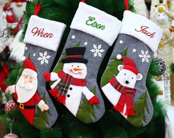May include: Three gray Christmas stockings with white cuffs hang on a green tree. Each stocking has a different design: Santa Claus, a snowman, and a polar bear. The names "Wren", "Etson", and "Jack" are embroidered on the cuffs.