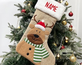 May include: A Christmas stocking with a reindeer design. The stocking is tan with a white cuff that reads "NAME" in red. The reindeer has a red and white hat, a green and white striped scarf, and brown antlers.