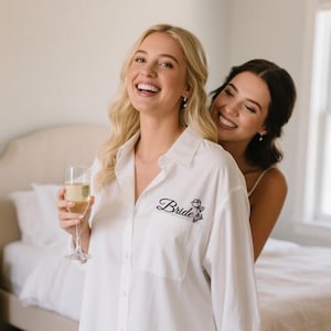 May include: A white button-down shirt with the word "Bride" embroidered in black script and a floral design. The shirt is worn by a person, with another person standing behind them. The setting is a bedroom.