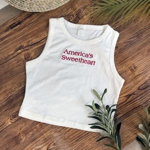 Può includere: Un top corto bianco senza maniche con scollo rotondo e la scritta America's Sweetheart ricamata in filo rosso sul petto. Il capo presenta un colletto a coste e un orlo dritto, caratterizzato da un design pulito ed essenziale.