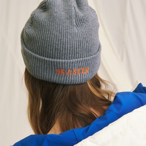 May include: Gray knitted beanie with the text "BE A SEE-ER" embroidered in orange on the back.