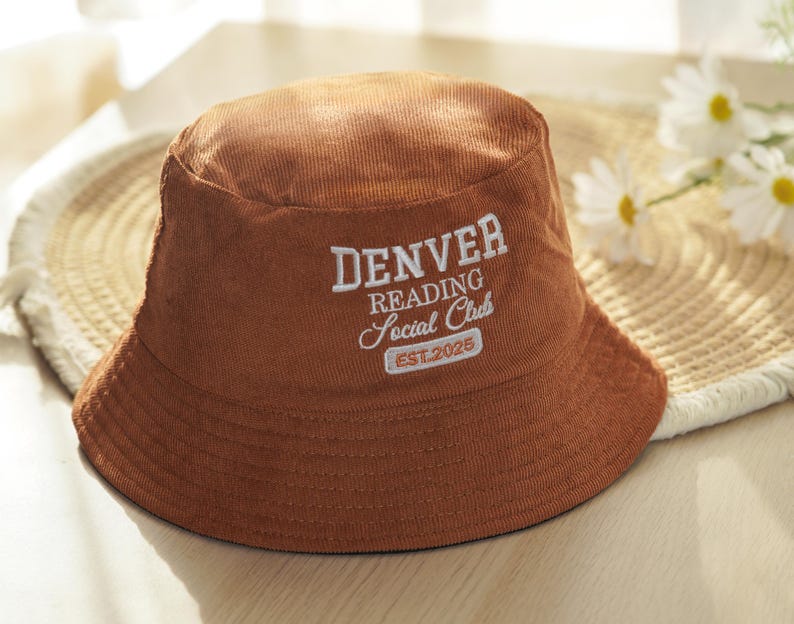 May include: A brown corduroy bucket hat with the text "DENVER READING Social Club EST.2025" embroidered in white. The hat is sitting on a woven mat with white flowers in the background. The hat has a wide brim.