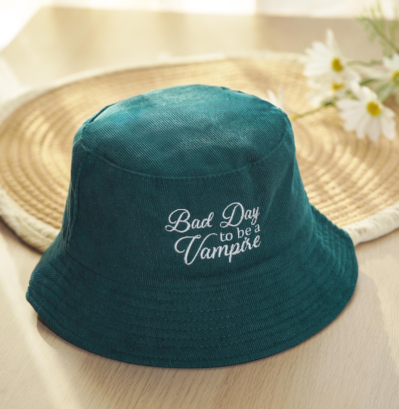 May include: A teal corduroy bucket hat with the text "Bad Day to be a Vampire" embroidered in white script. The hat is sitting on a woven straw surface.
