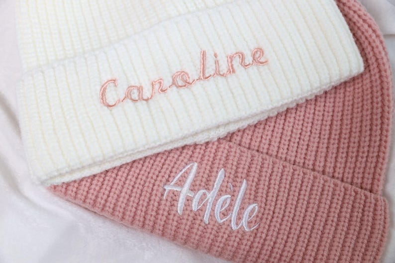 May include: Two knitted beanies, one white with the name "Caroline" embroidered in pink, and one pink with the name "Adele" embroidered in white.