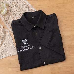 May include: Black button-down shirt featuring the "Blanco Fishing Club" logo embroidered in white. The shirt has a textured finish, a classic collar, and buttoned cuffs. It is displayed on a wooden surface.